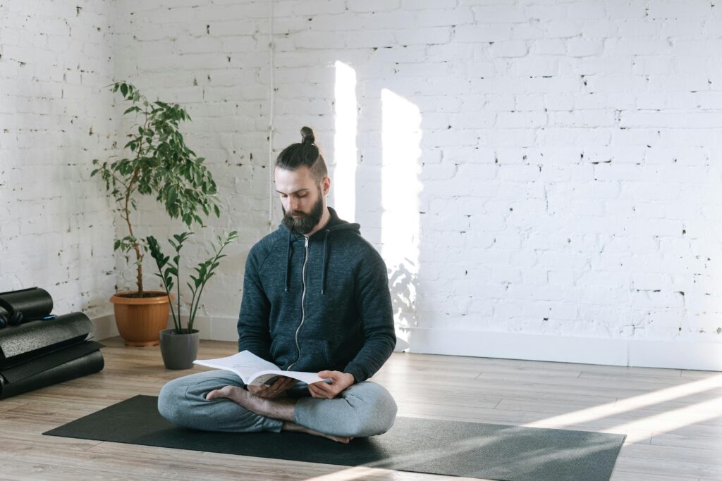 Man sitting cross-legged on a yoga mat in a bright, minimalist studio, reading a book with calm focus.