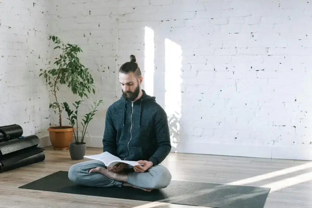 Man sitting cross-legged on a yoga mat in a bright, minimalist studio, reading a book with calm focus.